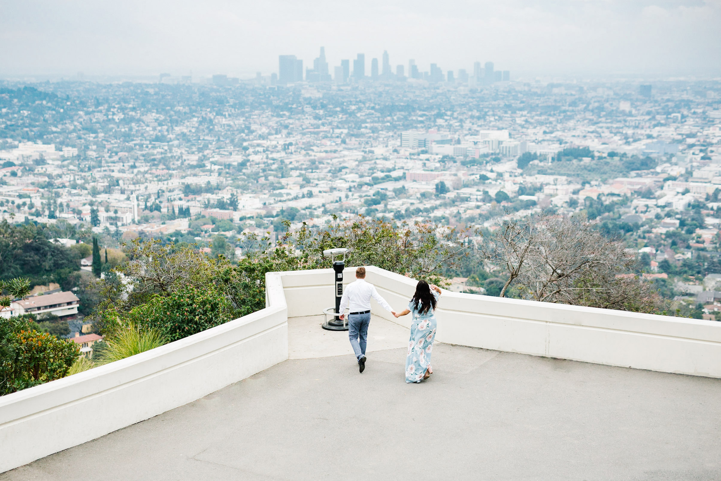 griffith observatory engagement photos Couple walking toward viewing scope at Griffith Observatory with the Los Angeles skyline in the background.